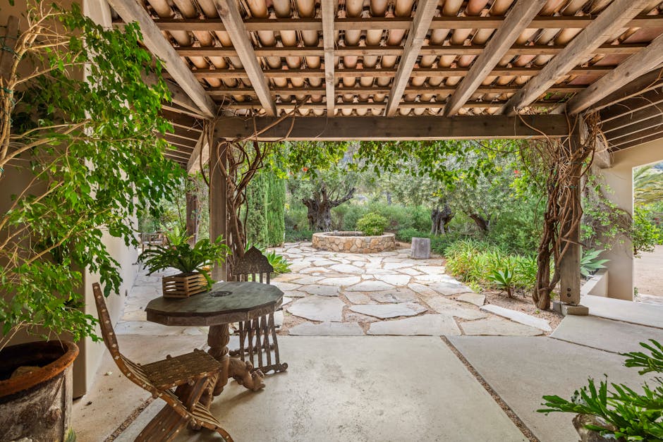 Rustic patio with wooden furniture, lush plants, and stone pathway in serene garden setting.